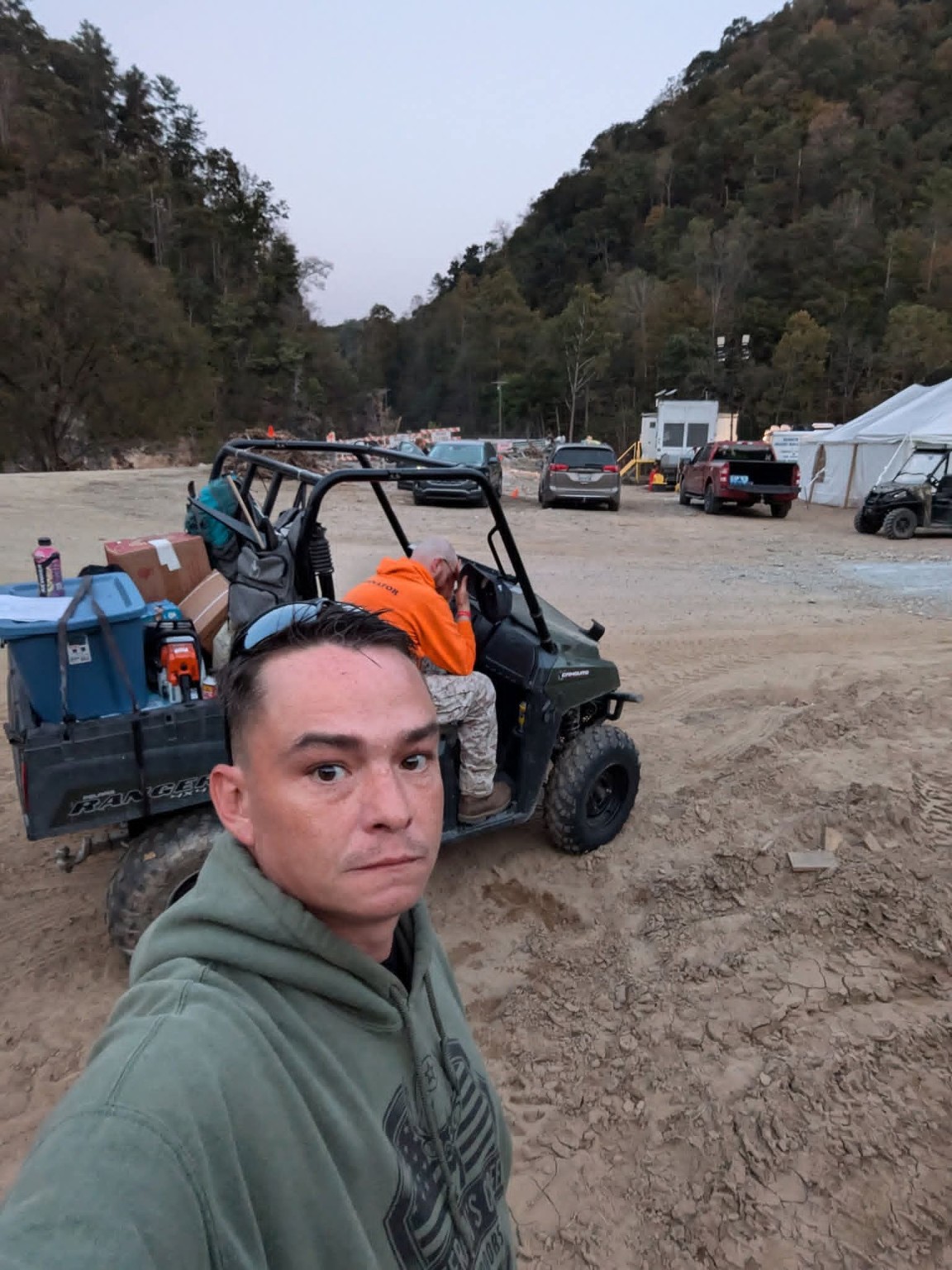 Ryan McClymonds at a mountain relief site with a utility vehicle, another worker, and the surrounding mountain community behind him.